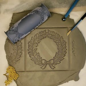 Puede incluir: Un rodillo de silicona gris con un dise&ntilde;o de corona y lazo. El rodillo se utiliza para crear una impresi&oacute;n en una pieza de arcilla. La arcilla tiene un dise&ntilde;o de corona y lazo impreso en ella.