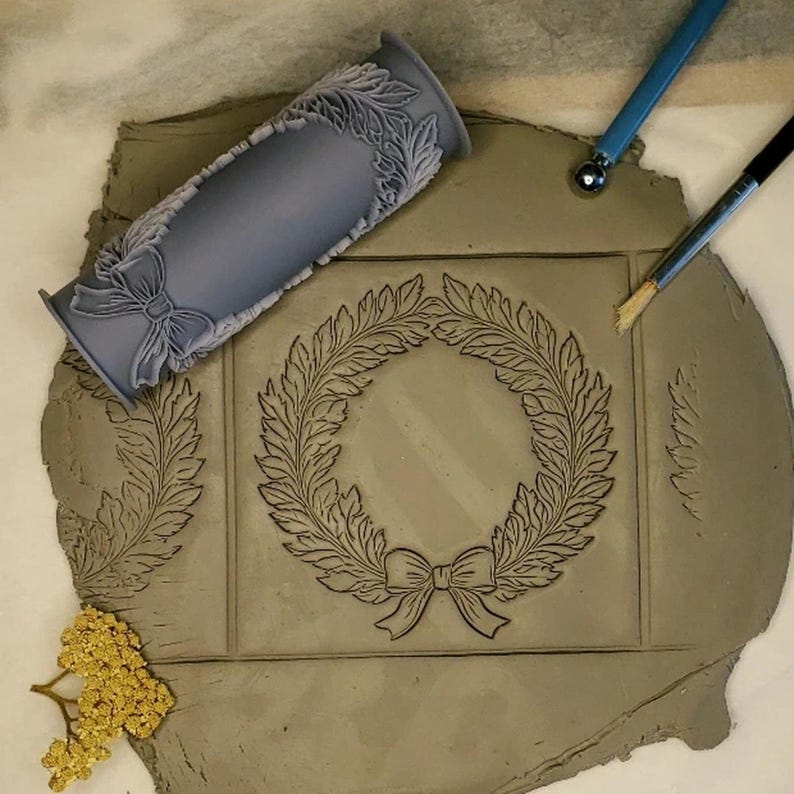 Puede incluir: Un rodillo de silicona gris con un dise&ntilde;o de corona y lazo. El rodillo se utiliza para crear una impresi&oacute;n en una pieza de arcilla. La arcilla tiene un dise&ntilde;o de corona y lazo impreso en ella.