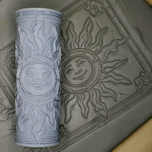 May include: A gray rolling pin with a raised design of a sun with a face. The sun has a circular frame with rays emanating from it. The rolling pin is next to a gray surface with a similar design.