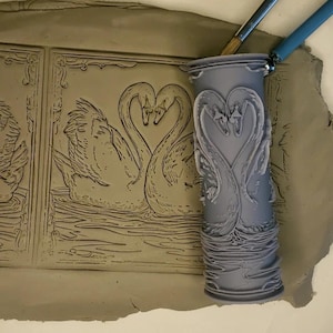 May include: A gray 3D printed rolling pin with a design of two swans forming a heart. The rolling pin is resting on a piece of clay with a similar design of two swans forming a heart.