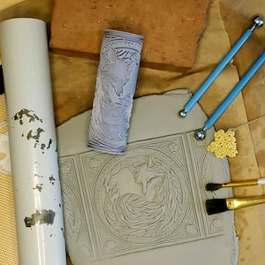 May include: A gray clay tile with a detailed design of two foxes curled up together. The tile is surrounded by a decorative border. A rolling pin with a fox design, two blue and silver tools, and a small pile of gold glitter are also visible.
