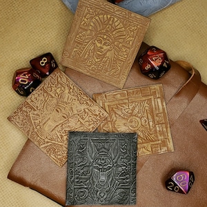 May include: Six brown leather coasters with intricate designs, including a winged creature, a stylized face, and a geometric pattern. The coasters are arranged on a brown leather surface with two red and one black dice.