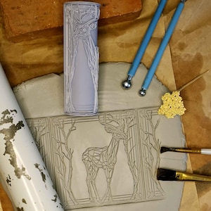 May include: A light purple cylindrical stamp with a deer design, alongside a clay impression of the same design. The deer is depicted in a geometric style, surrounded by trees. Additional tools and materials are visible.