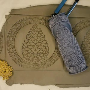 May include: A gray clay slab with a pine cone design imprinted on it. A gray rolling pin with a pine cone design is on the clay slab.