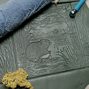 May include: A gray, textured clay slab with an embossed design of a beaver in a marsh setting. A roller with a similar design is in the upper left. A small cabin is visible in the background. A blue tool is in the upper right.