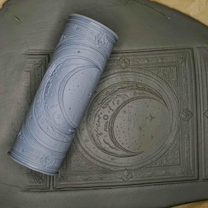 May include: A gray rolling pin with a crescent moon and stars design. The rolling pin is on a gray surface with a similar design.