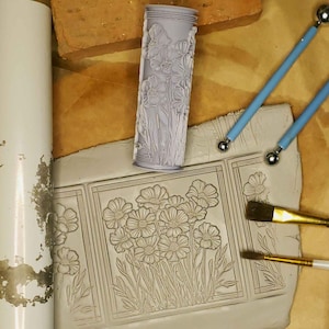 May include: A close-up of a clay tile with a floral design. The tile is surrounded by tools used for creating pottery, including a rolling pin, a brush, and a stylus.