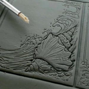May include: A detailed gray clay relief sculpture of a seashell with waves crashing around it.