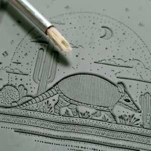 May include: A gray ceramic tile with an embossed design of an armadillo, cactus, and a crescent moon. The tile is being painted with a brush.