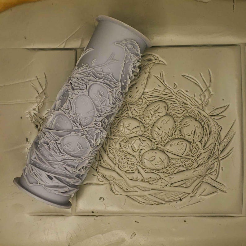 K&ouml;nnte beinhalten: Nahaufnahme eines grauen Gummibals mit einem erhabenen Design eines Vogelnests mit Eiern. Der Roller befindet sich neben einem Tonabdruck des gleichen Designs.