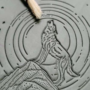 May include: A gray clay relief sculpture of a wolf howling at the moon. The wolf is standing on a mountain with a circular pattern behind it.