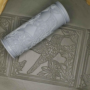 May include: A gray rolling pin with a hummingbird and floral design. The rolling pin is used to create impressions in clay or dough.