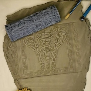 May include: A gray rolling pin with an elephant design is shown on a brown clay surface. The rolling pin is used to create an embossed elephant design on the clay.