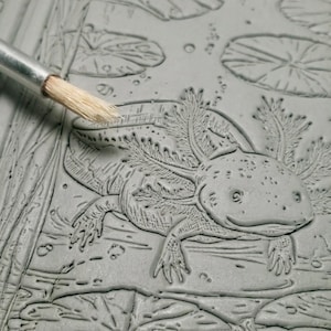 May include: A close-up of a gray relief carving of an axolotl in a pond setting, with a brush touching the surface. The axolotl is detailed with gills, feathery appendages, and a smiling expression. Lily pads and bubbles are also carved into the surface.
