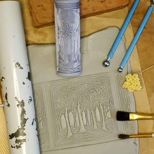 May include: A close-up of a clay slab with a design of trees and swirls. The design was created using a rolling pin with a similar design. There are also two small tools and a paintbrush on the table.