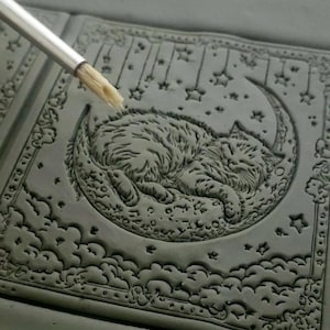 May include: A close-up of a gray clay tile with a detailed design of a cat sleeping on a crescent moon. The design is surrounded by stars and clouds. A paintbrush is being used to apply paint to the tile.