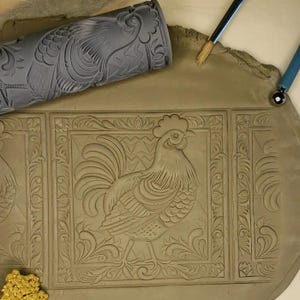 May include: A close-up of a clay surface with a rooster design imprinted using a rolling pin. The design features intricate details and a decorative border.