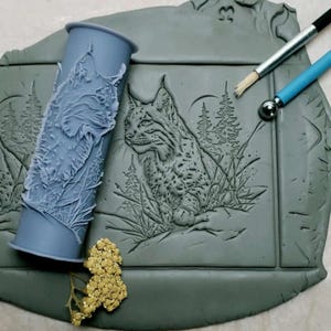 May include: A gray silicone rolling pin with a detailed design of a lynx in a forest setting. The rolling pin is next to a gray clay tile with the same design, but in a raised relief.