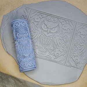 May include: A blue rolling pin with an owl design, used for creating embossed patterns in clay or dough. The rolling pin is shown next to a piece of gray clay with an embossed owl design.