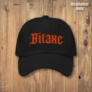 May include: Black baseball cap with the word "Bitaxe" embroidered in orange gothic lettering. The hat has a curved brim and a rounded crown. The background is a wooden surface.