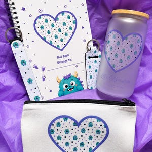 May include: A spiral-bound notebook with a heart-shaped monster pattern, a frosted glass tumbler with a bamboo lid, two fabric keychains, and a white canvas zipper pouch. Each item features a matching blue monster print and heart motif.