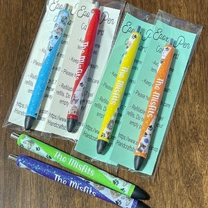 May include: Five colorful gel pens with black ink and a clear plastic cap. Each pen has a different design featuring a dog with a glitter finish. The pens are labeled "The Misfits" and have a website address on the packaging.