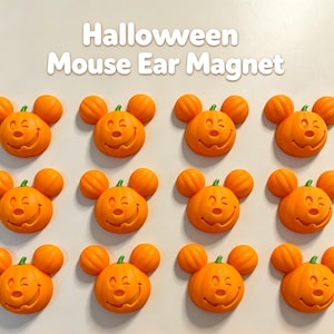 Puede incluir: Una colección de imanes naranjas con forma de calabaza y orejas de ratón, cada uno con una cara guiñando un ojo y una boca sonriente. El texto 'Halloween Mouse Ear Magnet' aparece en la parte superior sobre la cuadrícula de doce imanes.