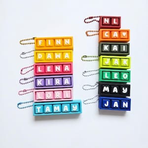 May include: Colorful keychain name tags in various colors, including orange, yellow, pink, purple, and blue. Each tag features a name in white letters. The names include Finn, Dawa, Lena, Kira, Lori, Tamay, NL, Cav, Kai, Jak, Leo, May, and Jan.