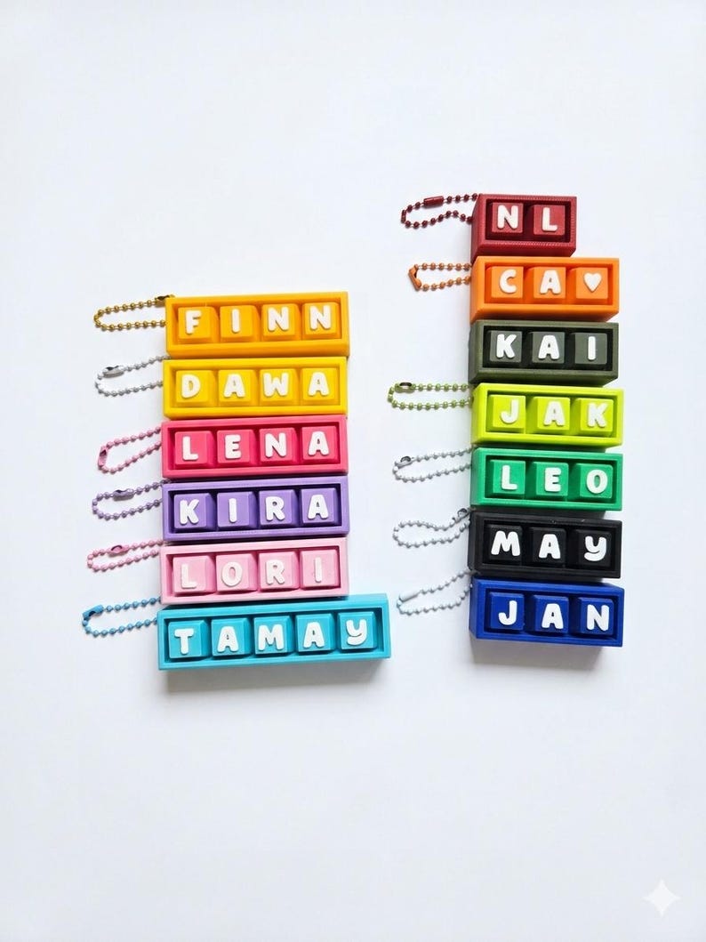May include: Colorful keychain name tags in various colors, including orange, yellow, pink, purple, and blue. Each tag features a name in white letters. The names include Finn, Dawa, Lena, Kira, Lori, Tamay, NL, Cav, Kai, Jak, Leo, May, and Jan.