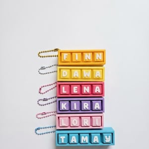 May include: A stack of colorful keychains, each with a different name spelled out in white letters. The keychains are in shades of yellow, red, pink, purple, and blue. Each keychain has a metal chain attached.