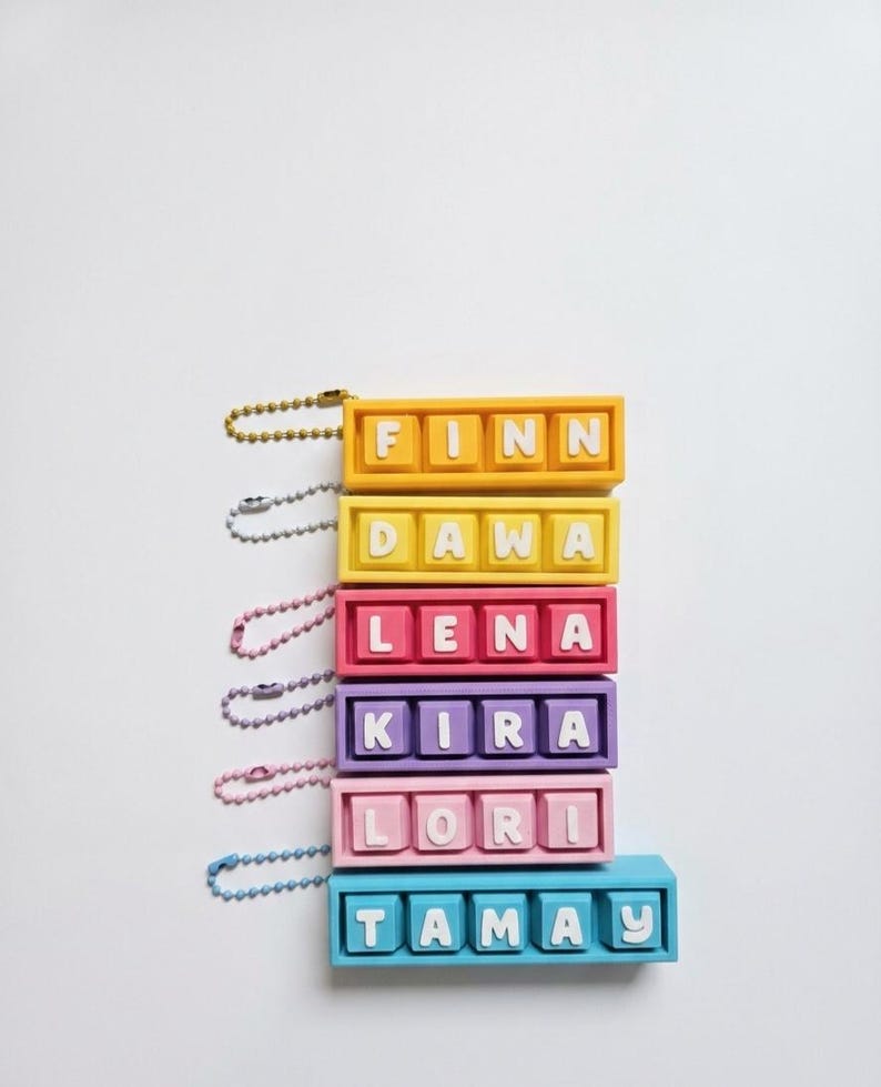 May include: A stack of colorful keychains, each with a different name spelled out in white letters. The keychains are in shades of yellow, red, pink, purple, and blue. Each keychain has a metal chain attached.