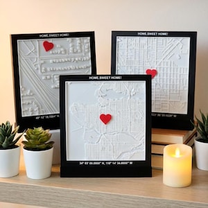 May include: Three framed 3D city maps with a white relief design and black frames. Each map has a red heart and the text "HOME, SWEET HOME!". One map has the coordinates 49° 12' 18.1584" N, another 34° 03' 08.0000" N, 118° 14' 34.0000" W, and the last 123° 06' 50.0820" W.
