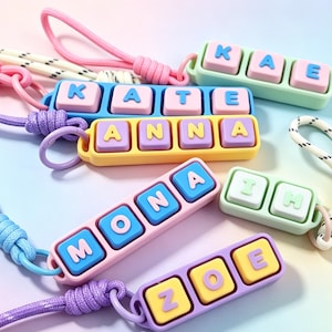 May include: A collection of colorful name keychains. Each keychain features a name spelled out with individual letter blocks in various colors, including blue, pink, yellow, and green. The keychains are attached to matching colored lanyards.