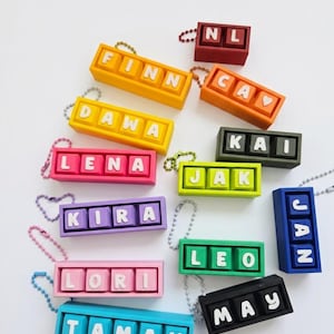 May include: A selection of colourful name keychains. Each keychain displays a name in white letters on a rectangular block. Keychains are in various colours, including yellow, orange, pink, purple, green, blue, and black. Each keychain has a small metal chain.