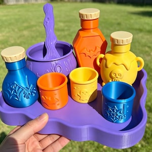 Peut inclure: Un plateau violet contenant des objets sur le thème d'Halloween, dont un chaudron violet avec un agitateur, une bouteille orange en forme de cercueil, un pot jaune avec un crâne, une bouteille bleue avec une toile d'araignée et trois petits gobelets.