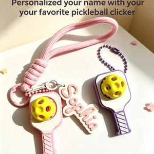 Puede incluir: Dos llaveros de paleta de pickleball con pelotas amarillas, uno rosa y otro morado, con una etiqueta blanca que dice Sophia. El llavero rosa tiene un cordón largo rosa, mientras que el morado tiene una cadena de cuentas. Texto: Personalized your name with your your favorite pickleball clicker.
