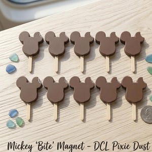 Puede incluir: Diez imanes marrones con forma de Mickey Mouse sobre palitos de madera, dispuestos en dos filas. Algunos imanes tienen un mordisco. El texto "Mickey 'Bite' Magnet - DCL Pixie Dust" está en la parte inferior.