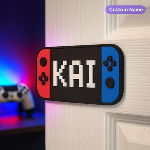 Puede incluir: Un letrero de puerta con forma de mando de consola de juegos, con secciones rojas, negras y azules. El nombre "KAI" se muestra en letras blancas pixeladas. Una etiqueta púrpura en la parte superior dice "Custom Name".
