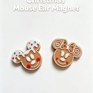 May include: Two gingerbread-style mouse ear magnets featuring white icing, red and green sprinkles, and red cheeks. The left magnet has white icing drips, while the right magnet includes a bow. The text Christmas Mouse Ear Magnet is above.