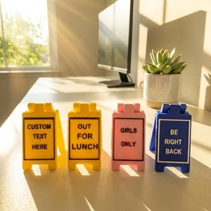 May include: Four small, colourful signs with text on a white desk. The signs are yellow, pink, and blue. The yellow sign says "CUSTOM TEXT HERE" and "OUT FOR LUNCH". The pink sign says "GIRLS ONLY". The blue sign says "BE RIGHT BACK".