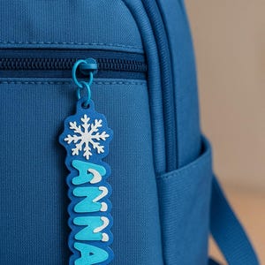 May include: A blue backpack with a matching blue zipper and a blue Anna keychain. The keychain features a snowflake design and the name "ANNA" in white and blue letters. The backpack has a small pocket.