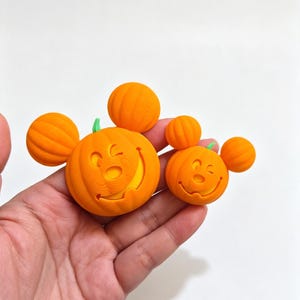May include: Two orange pumpkin-shaped ornaments with round ears and a carved face featuring a winking expression. Each pumpkin has a small green stem on top. The surface of the ornaments shows a textured, layered finish from the 3D printing process.