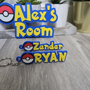 May include: Blue and yellow custom room sign that says "Alex's Room" with a cartoon design. Also, two keychains with the names "Zander" and "RYAN" in yellow letters on a blue background, with a cartoon design.