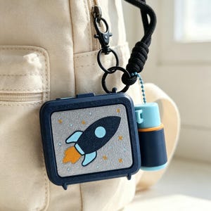 May include: A dark blue rectangular case featuring a space-themed graphic of a black rocket with orange flames and light blue accents against a gray background with yellow stars. A small matching blue and orange canister hangs from a black lanyard.
