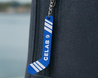 May include: A blue hockey stick keychain with white stripes at the top and bottom, featuring the white text "CELAB 9" printed vertically along the shaft. The keychain is attached to a silver metal ring and chain, hanging from a dark fabric zipper.
