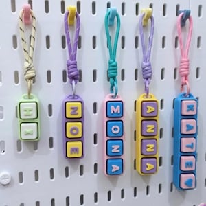 May include: Five colorful keychains with names spelled out in block letters. Each keychain has a different color scheme, including green, purple, pink, yellow, and blue. The names are Zoe, Mona, Anna, Kate, and a keychain with the letters H and R.