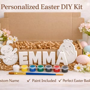 Op de afbeelding: Een gepersonaliseerde Paas DIY-kit met de naam "EMMA" in witte letters. De kit bevat houten uitsparingen in de vorm van een konijn en een kuiken, verfpotten, een penseel en decoratieve paaseieren. De kit wordt gepresenteerd in een kartonnen doos.