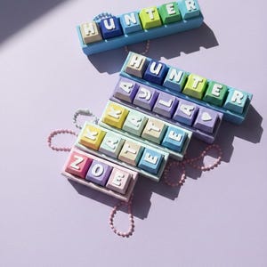 Op de afbeelding: Een verzameling kleurrijke, gepersonaliseerde naam sleutelhangers. Elke sleutelhanger toont een naam die is gespeld met individuele, pastelkleurige toetsenbordtoetsen. De namen "HUNTER", "ADLAE", "KATIE" en "ZOE" zijn zichtbaar. Een delicate roze kralenketting is aan sommige sleutelhangers bevestigd.