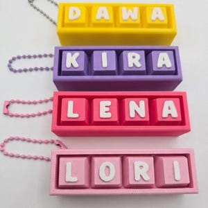 May include: Four colorful keychains with names: Dawa (yellow), Kira (purple), Lena (pink), and Lori (light pink). Each keychain has a matching chain. The names are in white block letters on square keys.