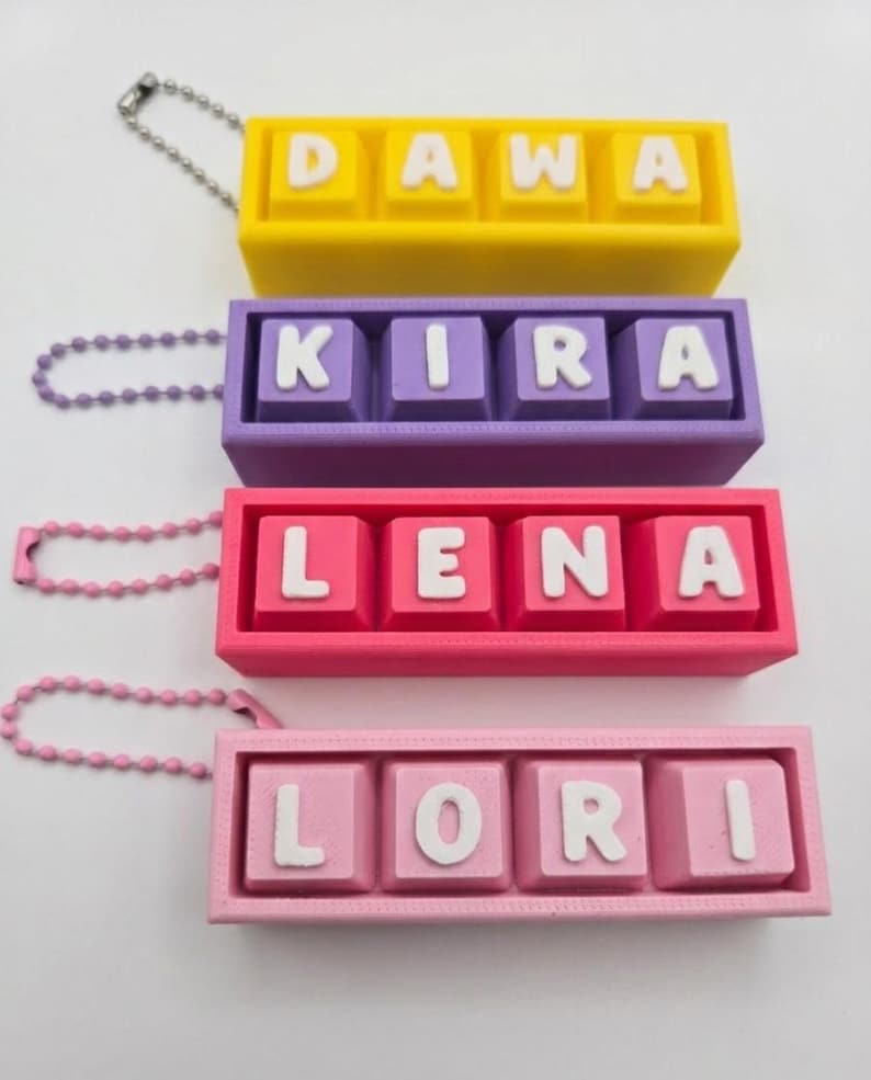 May include: Four colorful keychains with names: Dawa (yellow), Kira (purple), Lena (pink), and Lori (light pink). Each keychain has a matching chain. The names are in white block letters on square keys.